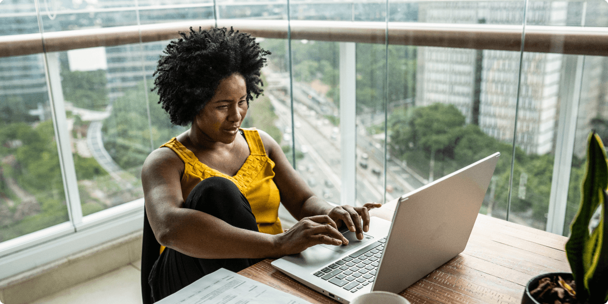 A woman working on her laptop in her apartment balcony. There is a view of trees and high-rise apartments behind her.