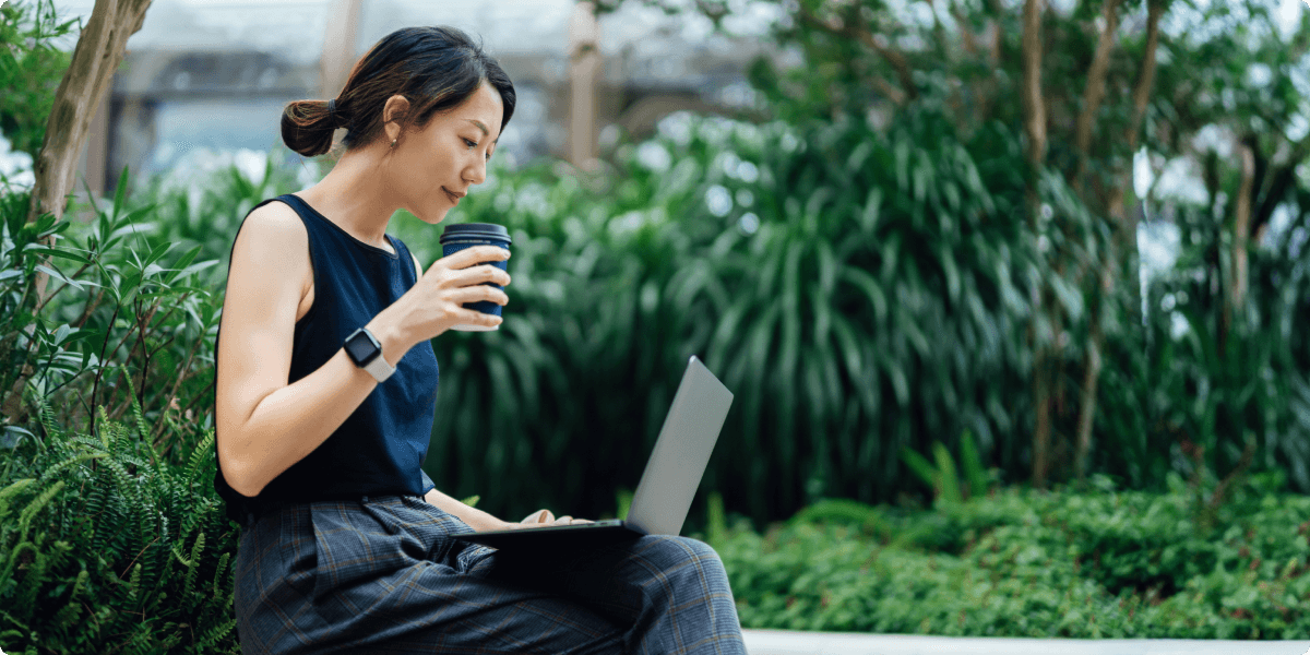 Una mujer en su portátil fuera con una taza de café. Está alrededor de vegetación exuberante.