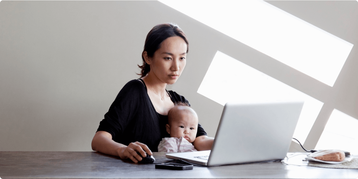 Une mère avec son enfant en bas âge sur les genoux, travaillant sur un ordinateur portable. La lumière du soleil passe derrière elle.