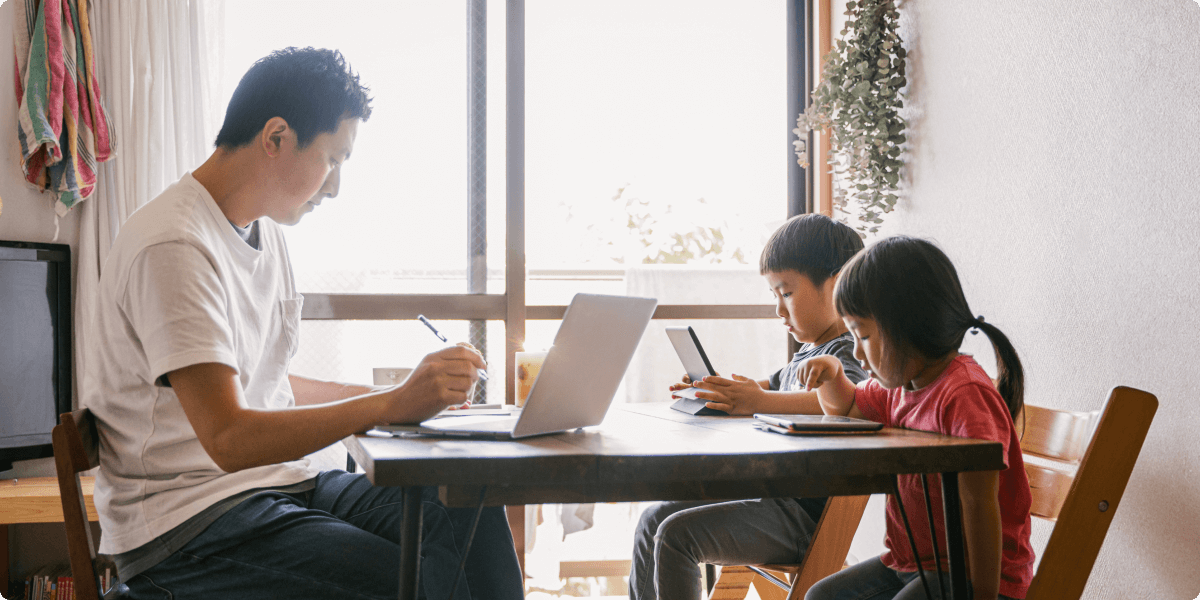 Um pai em casa, estudando com seus dois filhos na mesa da sala de jantar.